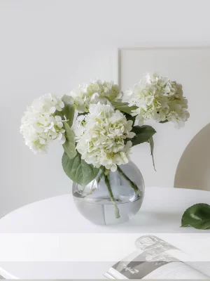 A stunning arrangement of four white artificial hydrangeas showcased in a clear glass vase. This arrangement brings timeless beauty and a refreshing aesthetic, perfect for any home or office space.