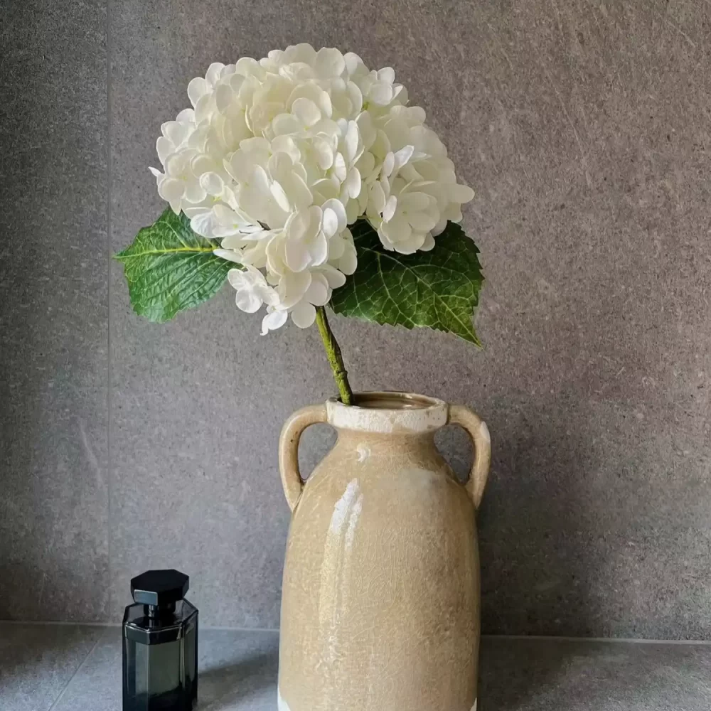 A single stem of white artificial hydrangea beautifully displayed in a cream-colored ceramic vase, adding a simple yet elegant touch to any decor with its lifelike petals and natural form.