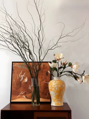 This artistic display combines faux magnolia flowers with a cream-yellow vase and another artificial branch, creating a vibrant and elegant setting. Set against the backdrop of a classical oil painting, the scene is full of life and texture, bringing a touch of nature into a richly decorated space.