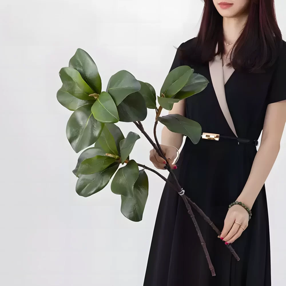 A side-by-side comparison of two artificial magnolia leaf stems, held by hand, showcasing the differences in length. One measures 23 inches and the other 28 inches, providing options for versatile floral designs.