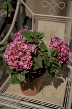 A vibrant faux pink hydrangea placed on a rustic outdoor chair, basking in warm sunlight. This arrangement adds a lively and refreshing touch to any outdoor space, perfect for patios or garden settings.