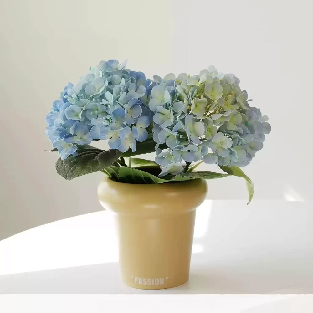 This small faux hydrangea arrangement features two blue hydrangea stems, elegantly placed in a cream-colored pot. The vibrant blue blossoms provide a charming and refreshing accent to any space, perfect for adding a pop of color to shelves or side tables.