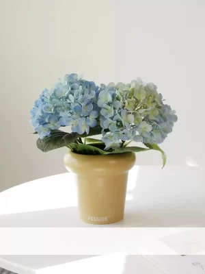 This small faux hydrangea arrangement features two blue hydrangea stems, elegantly placed in a cream-colored pot. The vibrant blue blossoms provide a charming and refreshing accent to any space, perfect for adding a pop of color to shelves or side tables.