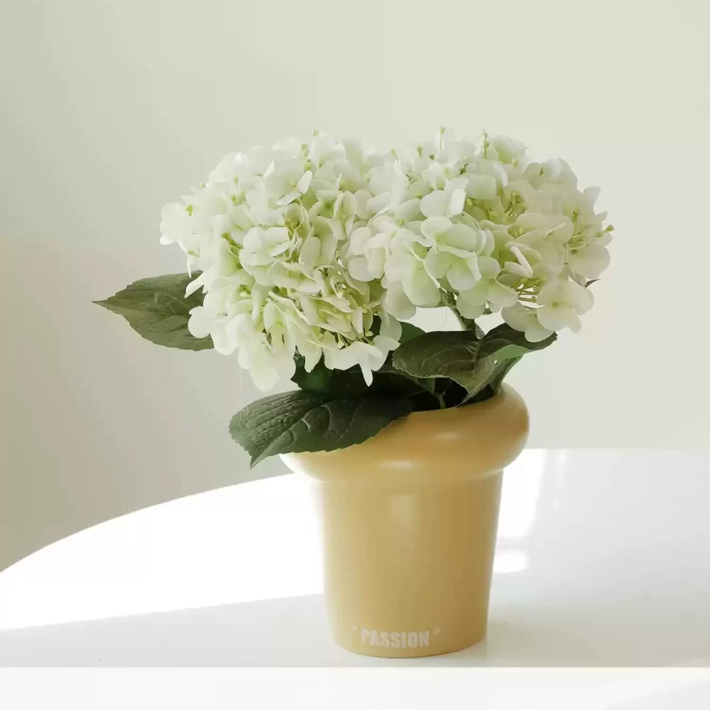 A minimalist display, this small artificial hydrangea arrangement includes two white stems in a stylish cream-colored pot. Ideal for brightening up any room with a touch of subtle elegance and sophistication.