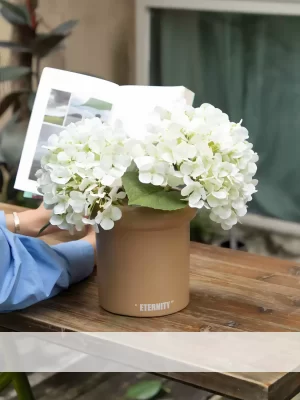 A serene and natural scene with a small faux white hydrangea arrangement, placed with books on a flower basket. Perfect for adding a touch of calm and elegance to any outdoor space.