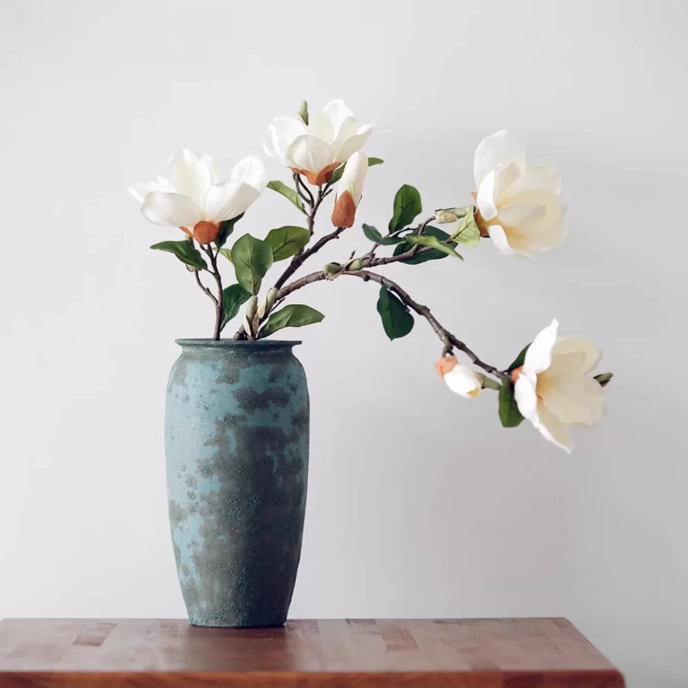 This arrangement features two realistic artificial magnolia stems, perfectly placed in a classic blue ceramic vase. The elegant vase complements the creamy magnolia petals, creating a stunning display that enhances any home decor or office setting.