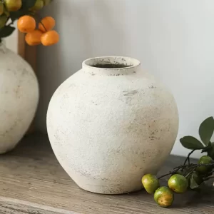 The vase sits comfortably on a table beside lifelike faux oranges, highlighting its organic appeal and earthy tones. The rounded design and textured finish add a touch of timeless elegance to any table or mantelpiece setting, bringing a cozy, rustic vibe to your home decor.